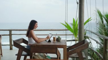 Kafede okyanus manzaralı, palmiye ağaçlı tablete resim çizen genç bir çizer. Oldukça odaklanmış bir kadın tropik kıyıların yakınındaki otantik bir barda çalışıyor. Freelancer Deniz kenarındaki illüstrasyon düzenleme.