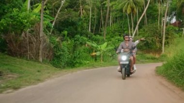 Motosiklete binen mutlu çift. İki sevgili tropiklerde motosikletle seyahat eder. Kız arkadaşı, erkek arkadaşı orman caddesinde scooter kullanıyor. Gezginler bisiklet yolculuğunun tadını çıkarırlar. Erkek, kadın macerada aşık.