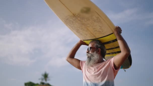 老年黑人男子将冲浪板举过头顶 以躲避太阳光 并准备在海水中冲浪 退休旅行时 戴着太阳镜的白胡子老人将独自在海滨游泳 — 图库视频影像