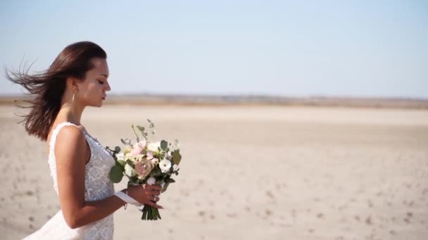 浪漫的新娘带着精致的婚礼花束在沙漠里 美丽女人的头发在大自然中随风飘扬 优雅的女士 白色的鲜花等待新郎 婚纱照 — 图库视频影像