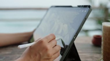 Serbest çalışan kadın eller tabletinde stilus ile resim çizer kafeteryada okyanus manzaralı buzlu kahve. Sahildeki bir restoranda grafik tasarımcısı çalışıyor. Sahilde uzaktan çalışan bir kadın. Yakın plan..