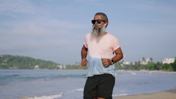 灰色のひげを持つ高齢のランナーマンは健康状態をチェックし 現代のフィットネストラッカーウォッチウェアラブルガジェットを使用し スポーツジョギング活動を行います 成熟した男性が熱帯の島と海のビーチに旅行 — ストック動画