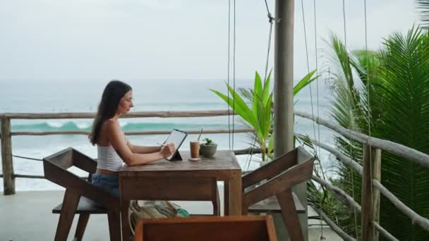 年轻的插画家在有海景 棕榈树的咖啡馆的石碑上画画 非常专注的女人在热带海岸附近的真正的酒吧里工作 海滨自由撰稿人编辑图解 — 图库视频影像