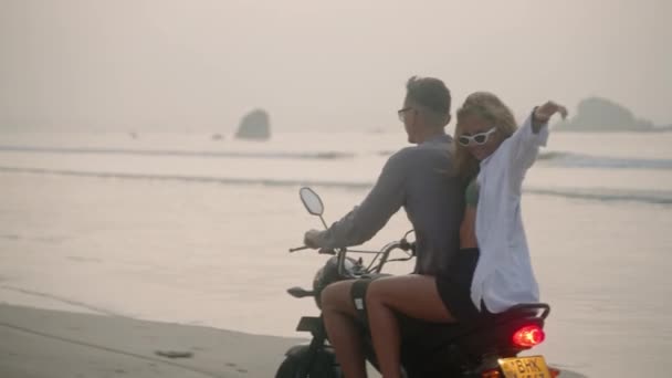 一对年轻夫妇在日落时骑摩托车在海滩上 女人骑摩托车沿着海岸线在海上旅行 女士转过头对着相机笑着张开胳膊在风中吹拂着头发 — 图库视频影像