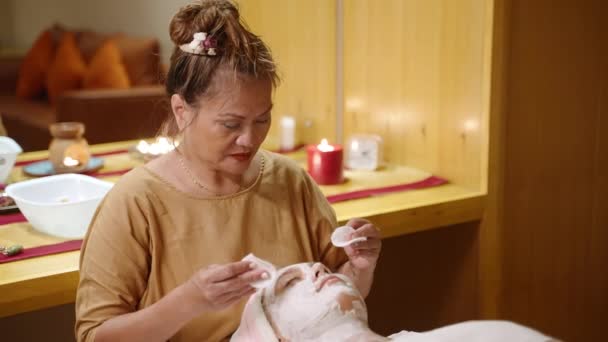 Kosmetolog Azjatycki Nakłada Białą Maskę Glinianą Twarz Klientki Centrum Odnowy — Wideo stockowe