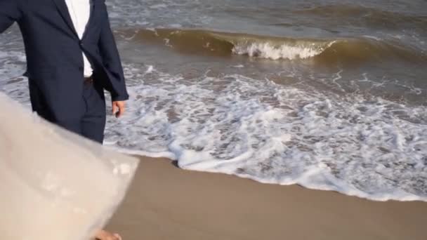 新郎的脚在沙滩上走着 婚礼花边裙在风中飘扬 男人的腿在海滩上慢慢地走 海上冲浪 恋爱中的快乐夫妻在海滨拍到了浪漫的照片 — 图库视频影像