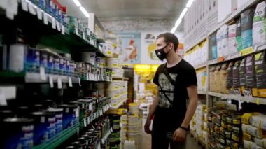 Yüz maskeli genç bir müşterinin ön tarafı hırdavatçıda koridorda yürüyor ve incelemek için raftan diş minesi alıyor. Bir adam DIY mağazasındaki mallara bakıyor.