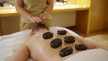 Kadın spa sağlık merkezi masajcısı müşteriye sıcak taşlar veriyor. Kadın masaj yapıyor, dinlenme tesisinde terapi yapıyor, yüzü yok. Kafkasyalı kişi kanepede dinleniyor. Sakinleştirici ağır çekim..