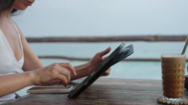Okyanus manzaralı kafede tablet üzerinde çalışan serbest çalışan kadın eller. Kadın tasarımcı deniz kenarındaki cihaza çizim yapıyor. Gezgin blogcu video blogu düzenliyor, kahve dükkanında cappuccino içiyor. Yakın plan, kenar görünüm.