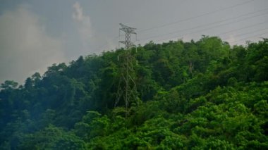 Yeşil yağmur ormanı kırsal kesimindeki elektrik hatlarının ve aktarım kulelerinin araba tarafındaki görüntüsü. Tepelik arazide elektrik dağıtım ağı var. Yüksek voltajlı güç kabloları. Bir yolculukta görünüm