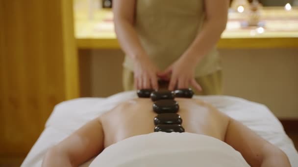 亚洲温泉健康中心的按摩师为病人的背部和颈部做滚石按摩 女人在放松工作室做热疗以减轻肌肉疼痛 白人没有脸的人在沙发上 — 图库视频影像