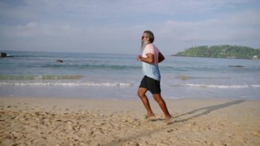 Gri sakallı yaşlı adam sabahları okyanus sahilinde modern fitness izleyicisiyle koşar kulaklıkla müzik dinler. Sağlıklı yaşlı siyah adam koşuyor, vücut gücünü arttırıyor ve spor yapıyor..