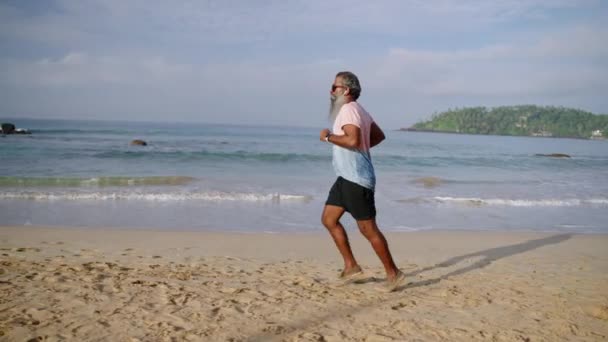 灰色のひげ付きの高齢男性は 海のビーチで午前中にモダンなフィットネストラッカーウォッチで走り イヤホンで音楽を聴きます 健康なシニアブラック老人ジョギング 体力を向上させ フィットネスをする — ストック動画