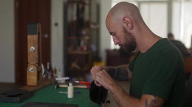 Genç profesyonel deri işçisinin yan görünüşü çalışma masasında oturur ve atölyede siyah el çantası yapar. Haki tişörtlü, sakallı, cesur bir adam küçük bir stüdyoda çanta yapıştırıyor..