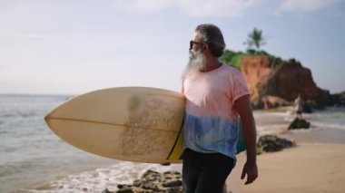 Kıdemli sörfçü sörf tahtası tutuyor ve tropik bir adada egzotik plajda yürüyor. Emekliliğinde güneş gözlüklü yaşlı zenci adam turizmin, okyanus manzarasının, gökyüzünün, güneş ışığının ve kayalık tepelerin tadını çıkarıyor..