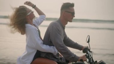 Güneş gözlüklü çift gün batımında deniz sahilinde motosiklet sürüyor. Mayo giyen kadın sarılan adam, rüzgar saçlarına dokunuyor, romantik bisiklet sürerken kameraya gülümsüyor. Kadın motorsikletle okyanusla seyahat ediyor..