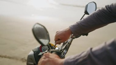 Motorcunun eli anahtarı motorun ateşlemesine yerleştiriyor, motoru çalıştırmak için okyanus kıyısında çalıştırıyor. Hız göstergesi olan bir motosiklet konsolu. Adam scooter 'ı çalıştırdı ve uzaklaştı. Yakın çekim.