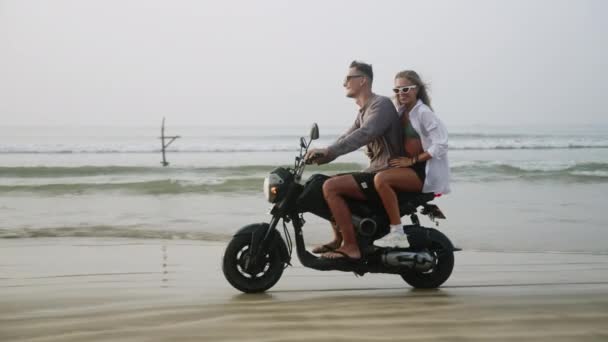 年轻夫妇骑摩托车 日落时在潮湿的沙滩上洒水 女人有乐趣驾驶摩托车在海洋海岸 女士大叫 张开双臂 拥抱男朋友 开始一段浪漫的旅程 — 图库视频影像