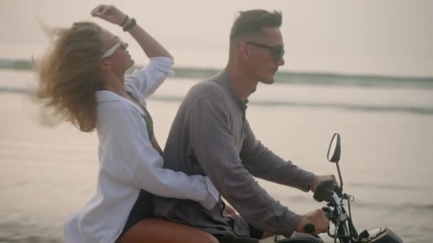 日落时 戴着太阳镜的情侣骑摩托车在海滩上 身着泳衣的女士拥抱着男人 触摸着风吹来的头发 在浪漫的自行车道上对着相机微笑 女人骑摩托车在海上旅行 — 图库视频影像