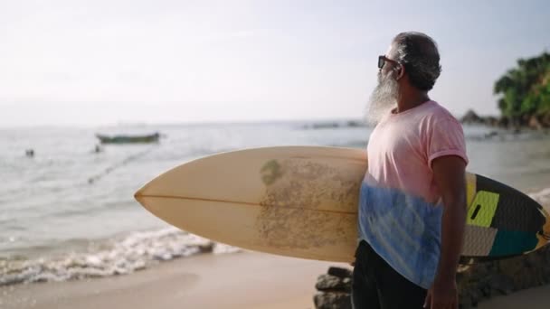 老年男性游客与冲浪板一起在海滩上散步 在黄金岁月有着积极而健康的生活方式 环顾四周 欣赏船湾风光 异国情调的自然和植物 海浪汹涌的海洋 — 图库视频影像