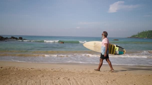 资深冲浪客手持冲浪板 在热带岛屿上奇异的海滩上散步 退休时戴太阳镜的黑胡子老人喜欢旅游 观赏大海 阳光和岩石山 — 图库视频影像