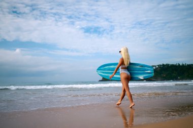 Deniz kenarında sörf tahtası olan genç bayan sörfçü sörf oturumuna hazırlanıyor, verimli yamaçlı tropikal plaj ortamı, aktif tatil takibi, sörf kültürü, doğal kıyı ortamında macera