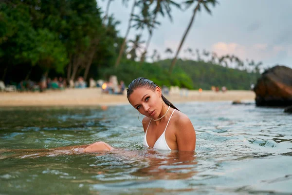 Female traveler swimming in tropical ocean, wears long-lasting makeup. Woman in stylish swimsuit takes a dip in sea. Vacationing near palm-lined beach, fashionable water-resistant cosmetics.