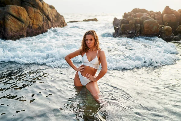 Beautiful woman poses in white bikini on seaside, rocks behind, wading in ocean water. Beautiful woman poses in white bikini on seaside, rocks behind, wading in ocean water.