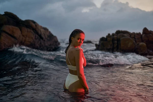 Elegant lady takes pleasure in late-night dip in sea, bathed in ruby luminescence. Fashionable swim attire on female, peaceful oceanic scene at night, serene aquatic environment.