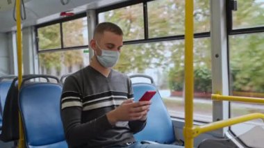 Genç erkek yolcu, tıbbi maske takarak tramvayla seyahat ediyor. Günlük kıyafetli bir adam otobüste oturur, akıllı telefon kullanır ve kişisel koruma malzemeleriyle pencereden dışarı bakar..