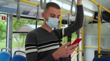 Tıbbi maske takan, akıllı telefon kullanan ve tırabzan taşıyan tramvayla seyahat eden genç bir yolcuyu yakından çek. Günlük giysili adam toplu taşımacılıkta COVid-19 'dan korunuyor..