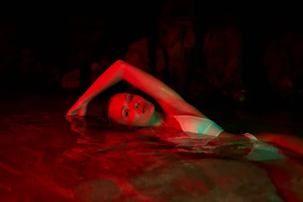 Female model swimming in night lagoon, red lighting. Beauty and femininity blend with natural pool setting. Creative aquatic photo session. Wet swimsuit on woman. Pose in water at dusk.