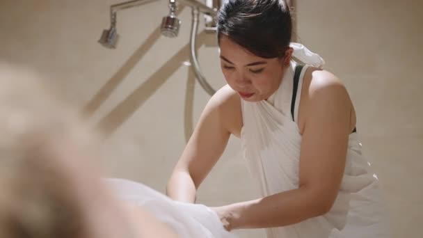 アジアのマッサージ師は スパハンマームで女性の足をこすりつぶしています サウナで洗浄 クレンジングによるアンチエイジングおよびアンチセルライト療法 女性はテーブルでリラックスして 全身の皮膚治療を受ける — ストック動画