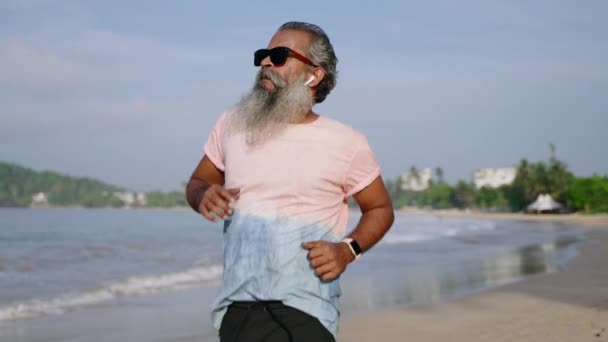 白胡子老人早上带着现代健身跟踪器在海滩上跑步 在耳边听音乐 健康的老年黑人男子慢跑 增强体格力量和健身 — 图库视频影像