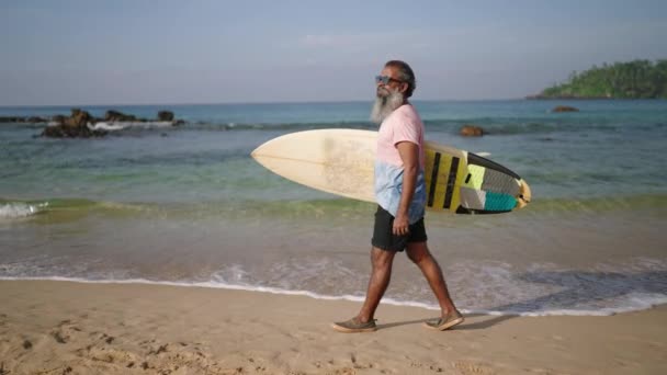 资深冲浪客手持冲浪板 在热带岛屿上奇异的海滩上散步 退休时戴太阳镜的黑胡子老人喜欢旅游 观赏大海 阳光和岩石山 — 图库视频影像