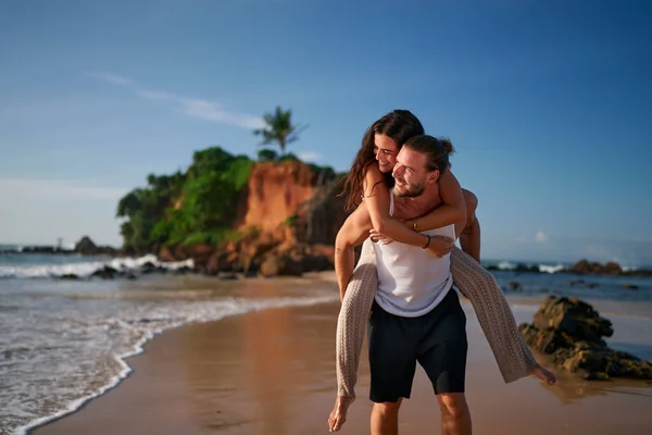Man carries woman piggyback, strolling along sea. Loving couple enjoys honeymoon on exotic beach. Carefree lovers embrace, walk on sand near ocean, cliff. Romantic getaway, unity, joy, love.