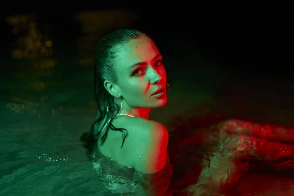 Female model submerged in reflective night waters, under green and red lighting, depicts contemporary glamour and chic in a fashionable night-time aquatic setting, portraying a serene ambiance.