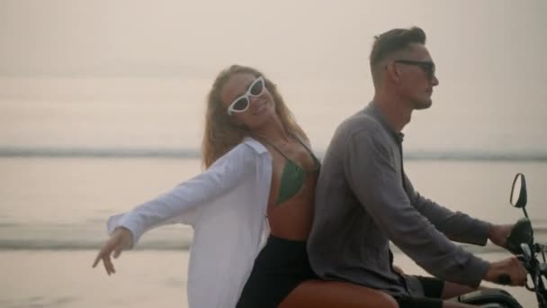 日落时 戴着太阳镜的情侣骑摩托车在海滩上 身着泳衣的女士拥抱着男人 触摸着风吹来的头发 在浪漫的自行车道上对着相机微笑 女人骑摩托车在海上旅行 — 图库视频影像