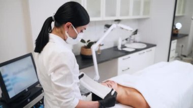 Kadın yaşlanma karşıtı prosedür için yatağa uzanıyor. Güzellik uzmanı, müşterisini modern klinikte cilt tedavisine hazırlıyor. Profesyonel olarak koruyucu saç bandı takar, deriyi temizler, hijyen ve bakım sağlar..