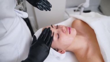 Kadın müvekkil yaşlanmayı önleme terapisinden hoşlanıyor, rahatça yatıyor. Profesyonel estetikçi cilt bakımı için yüz masajı yapar, kuaförde gençleştirme. Güzellik uzmanlarının elleri nemlendirilmiş cilt üzerinde çalışır, gözenekleri temizler..