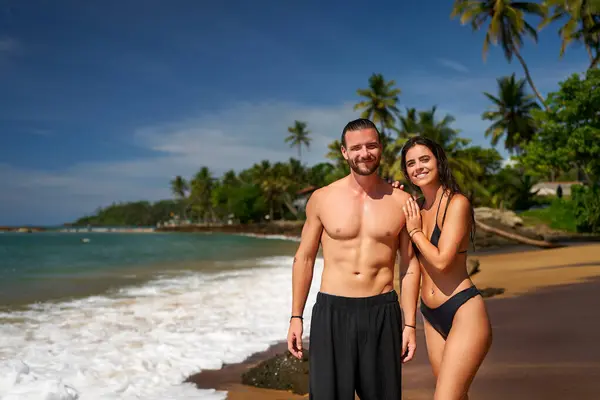 Abs, attractive duo on vacation, seaside workout, sunny coastal lifestyle, healthy living concept. Fit couple stands on tropical beach, athletic man and slender woman smile, enjoying eco travel.