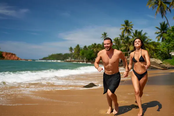 Couple in active lifestyle, summer vacation sprint. Cheerful man and woman enjoy beach run, barefoot by sea, palm trees background. Joyful jog, sunlit coast, ocean living freedom, fitness together.