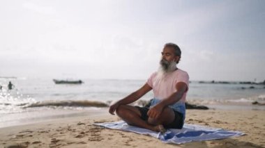 Yaşlı gri sakallı gezgin, zihnini boşaltmak için meditasyon ve yoga yapıyor, tropik adada lotus poz veriyor. Gözleri kapalı yaşlı bir adam, plajda palmiye ağaçları ve okyanusla emekliliğinin tadını çıkarıyor..