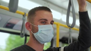 Tıbbi maske takmış, elinde tırabzan taşıyan genç bir yolcunun tramvay yolculuğu görüntülerini yakından çek. Günlük kıyafetler giyen Avrupalı adam toplu taşımacılıkta covid-19 'dan korunuyor.