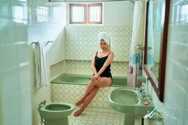 Self-care routine in classic style home spa. Woman relaxes in vintage bathroom, sitting by tub wearing swimwear, towel on head. Feminine leisure, wellness bath ritual, retro interior decor.
