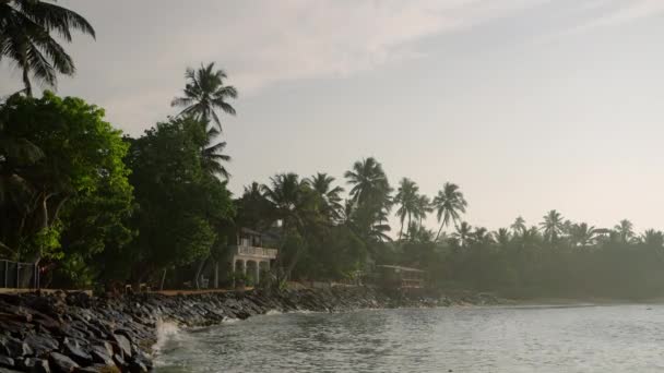 热带异国情调的海滩 有小旅馆和被绿树和棕榈树环绕的房子 海浪拍打在海滨的石头上 日落时的豪华度假胜地荒芜的海滨 前往温暖的国家 — 图库视频影像