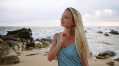 Dalgalar kayaları okşar, gün batımı yaklaşır, tatil atmosferi etrafını sarar. Gök mavisi elbiseli sarışın, dingin tropikal sahillerden hoşlanıyor. Boş zaman, özgürlük, kadın güzellikten zevk alır..