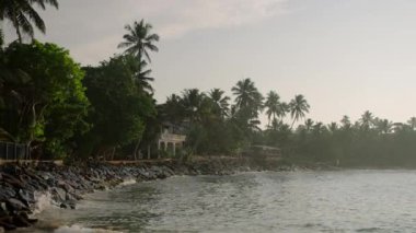 Yeşil ağaçlar ve palmiyelerle çevrili küçük otelleri ve evleri olan tropik egzotik plajlar. Deniz kıyısı taşlarına dalgalar sıçrıyor. Gün batımında lüks bir tatil köyünün ıssız sahillerinde. Sıcak ülkelere git..