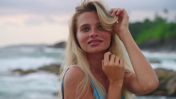 黄昏时穿着蓝色衣服的安详女士在海边 优雅的金发姑娘漫步在热带海岸 自然美 海浪背景 柔和的微笑 女性魅力 海滨度假视频 — 图库视频影像