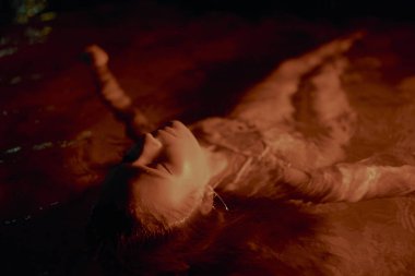 Kadın gece suda zarifçe uzanır, vücudu loş, sıcak bir ışıkla yıkanır, sakin, sakin bir sahne yaratır. Kadınlığın fotoğrafı, cazibe, doğal element güzelliği, yaratıcı sanat anlayışı..